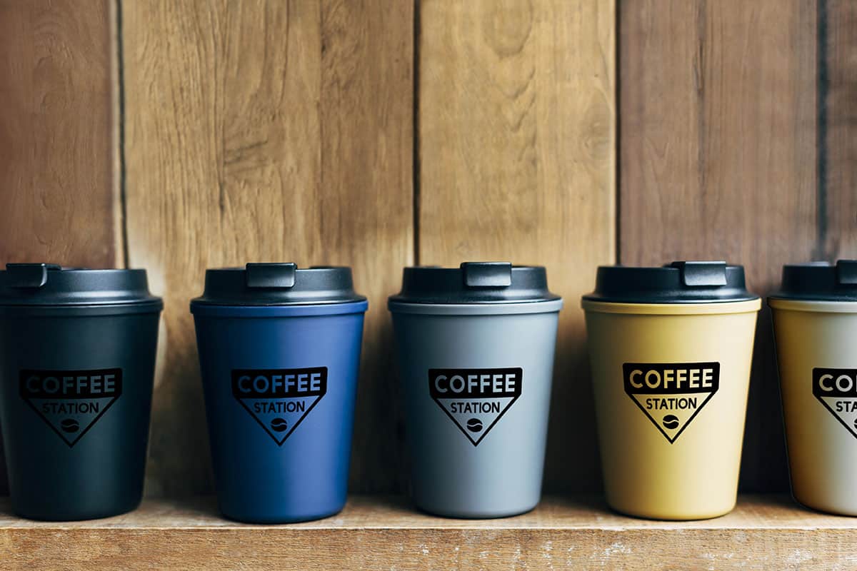 Branded Tumblers A row of four coffee cups in varying colors—black, blue, gray, and yellow—each with a black logo that reads "COFFEE STATION," set against a wooden background.